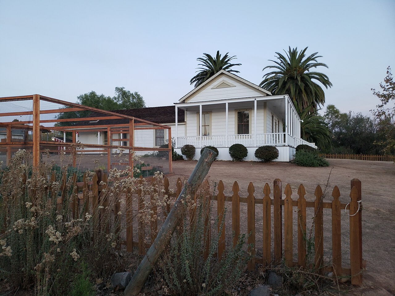 White historic farmhouse at Sikes Adobe Historic Farmstead seen behind a wooden fence and garden plantings.